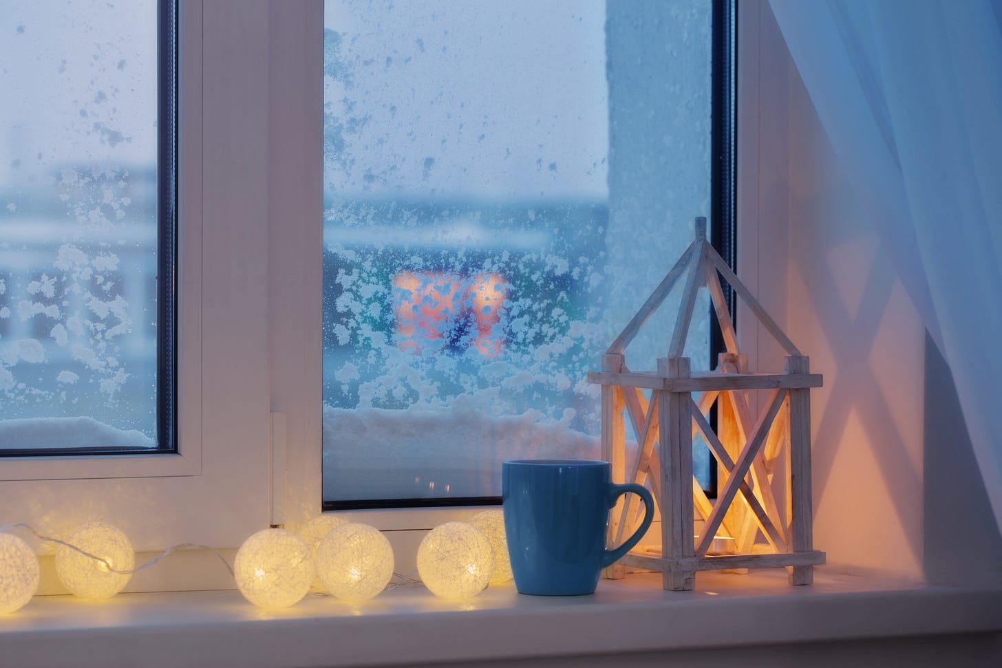 Close-up of a white windowsill. There’s a wooden lantern and a string of white balls, glowing softly, next to a blue mug. There’s snow accumulated in the outside of the window, and a few snowflakes cling to the windowpane. In the distance another window is illuminated by soft, yellow light.  