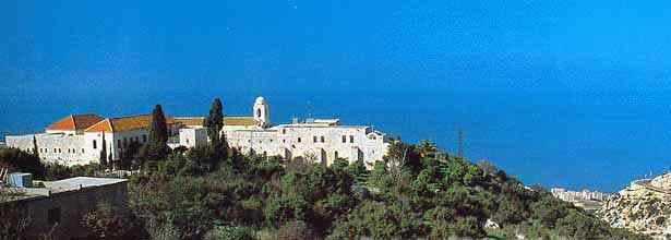 Panoramic view of a white monastery complex with a bell tower on a green hillside above the sea.