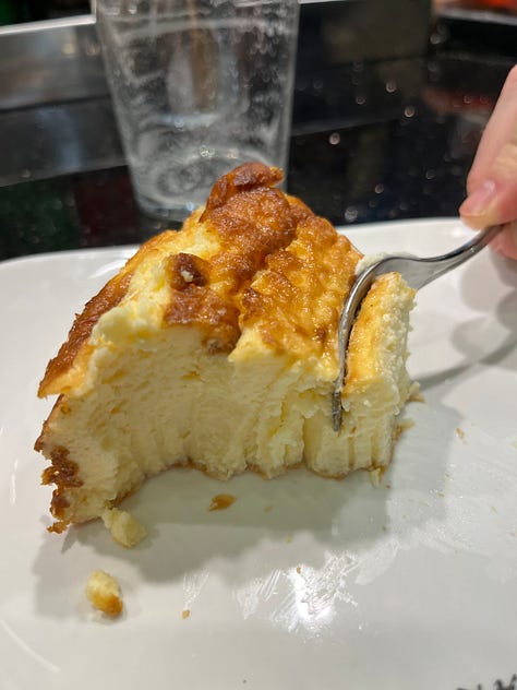 Crab stuffed squid, a spoon entering creamed uni in its shell, a hand holding a sesame crack with three different color pieces of smoked fish on top , a hand holding grilled foie on bread, a hand holding txistorra skewered onto a piece of bread, a fork entering a half eaten piece of cheesecake
