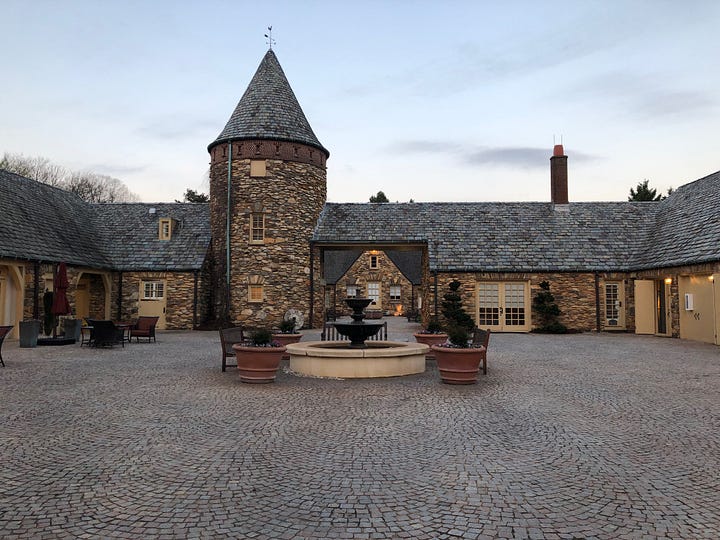 Two stone buildings in mock medieval style, each with a tower with a pointed top. The one on the left faces a lawn. The one of the right, a gravel courtyard with a fountain in the middle.