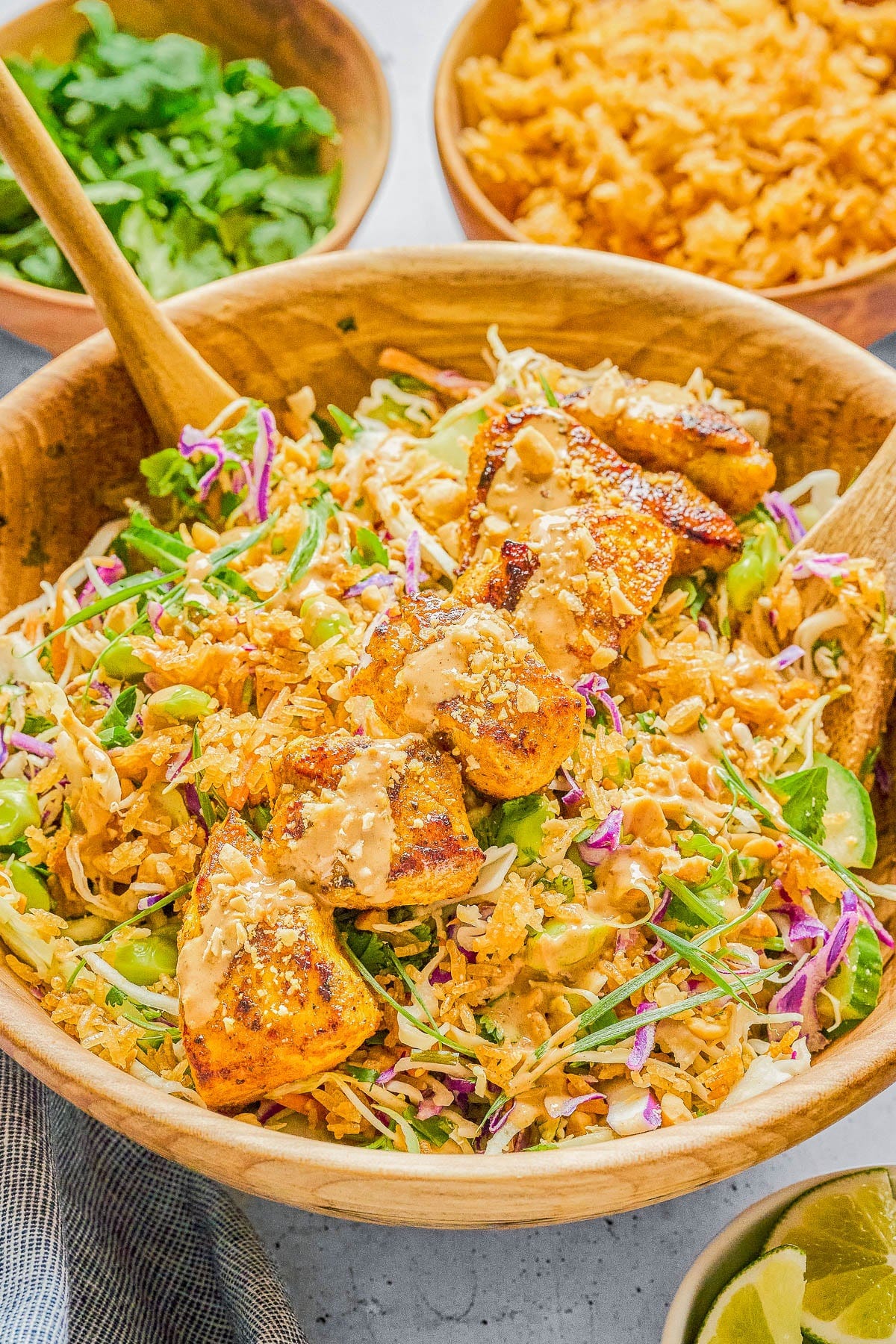 A wooden bowl filled with salad greens, shredded cabbage, grilled tofu, peanuts, and a creamy dressing, with wooden salad servers and side bowls of rice, cilantro, and lime wedges. A wooden bowl filled with salad greens, shredded cabbage, grilled tofu, peanuts, and a creamy dressing, with wooden salad servers and side bowls of rice, cilantro, and lime wedges.