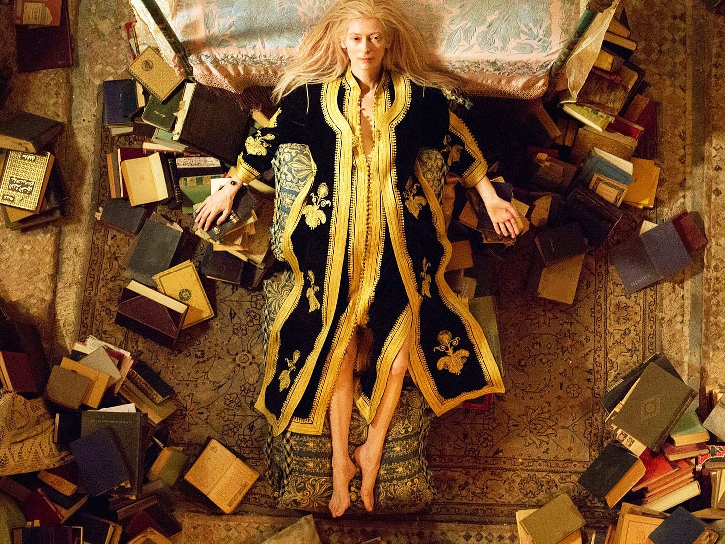 cena vista de cima. Tilda Swinton é uma mulher muito branca e loira, com os cabelos loiros longos e desarrumados. Ela está deitada no chão, com a cabeça encostada na cama. Ela é o centro da cena, cercada por livros espalhados por todo o recinto. Ela está em cima de um lindo tapete cheio de adornos, e usa um roupão ou quimono, de tecido escuro e detalhes em dourado, com motivos marroquinos