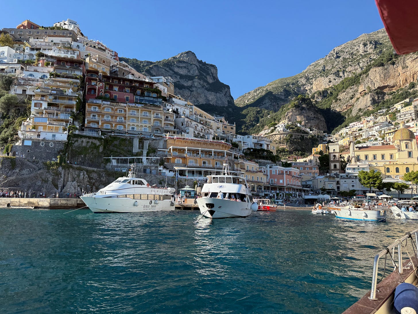 View of Positano's colorful cliffside buildings from the water, with boats anchored in the foreground and bright blue sky above. View of Positano's colorful cliffside buildings from the water, with boats anchored in the foreground and bright blue sky above.