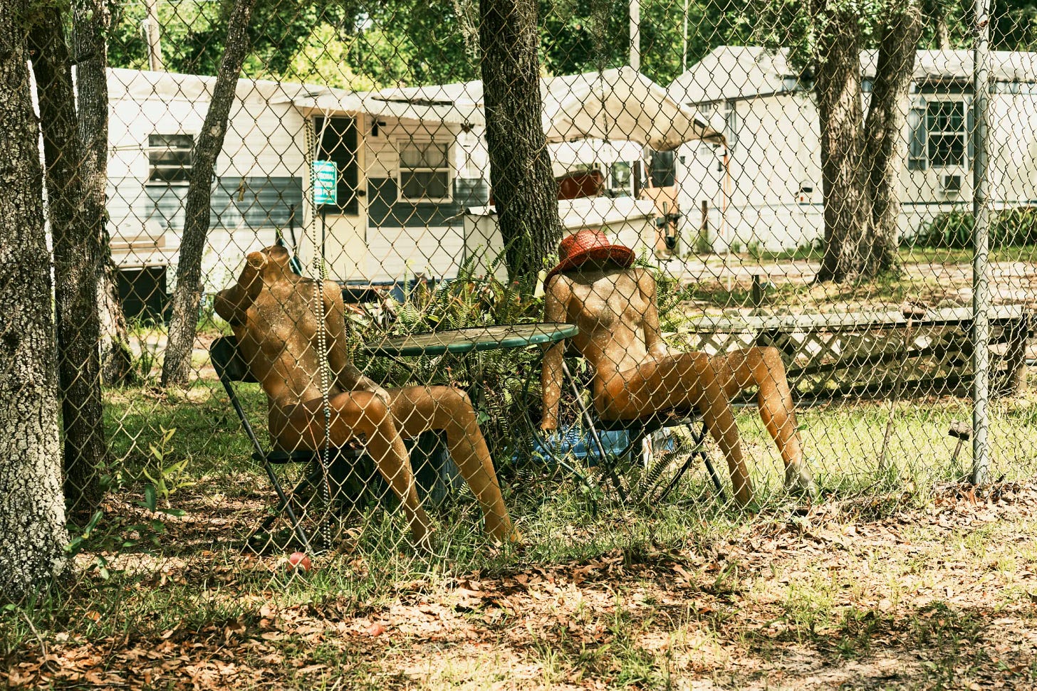 Two nude mannequins seated at a small outdoor table behind a chain-link fence, with mobile homes and trees in the background at Florida Naturist Park.