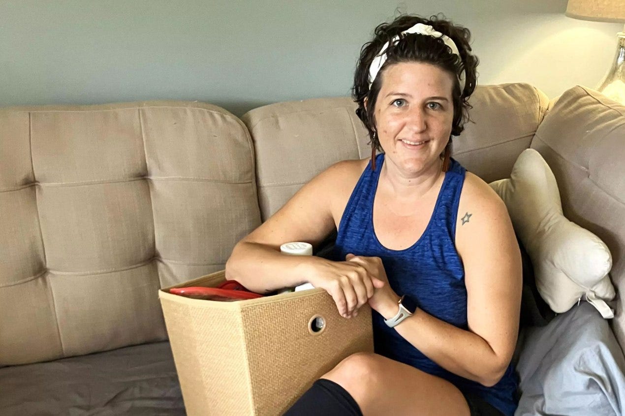 A woman with short brown hair held back with a white headband sits on a beige couch with a box in her lap filled with things. She is wearing long earrings and a blue tank top. Her legs are crossed and she is wearing knee-high black socks. A woman with short brown hair held back with a white headband sits on a beige couch with a box in her lap filled with things. She is wearing long earrings and a blue tank top. Her legs are crossed and she is wearing knee-high black socks.