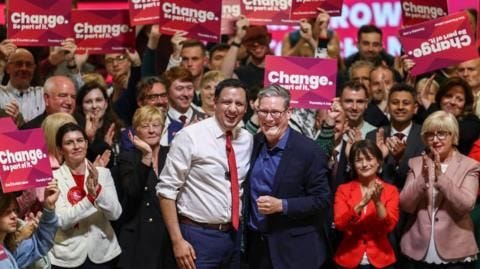Scottish Labour leader Anas Sarwar (a man with dark hair and a red tie, in a white shirt) posing with Sir Keir Starmer (a man with greying hair and glasses, wearing a dark suit with a blue shirt, no tie) at a Labour party rally - everyone is clapping and smiling, and many are holding up placards reading "change".