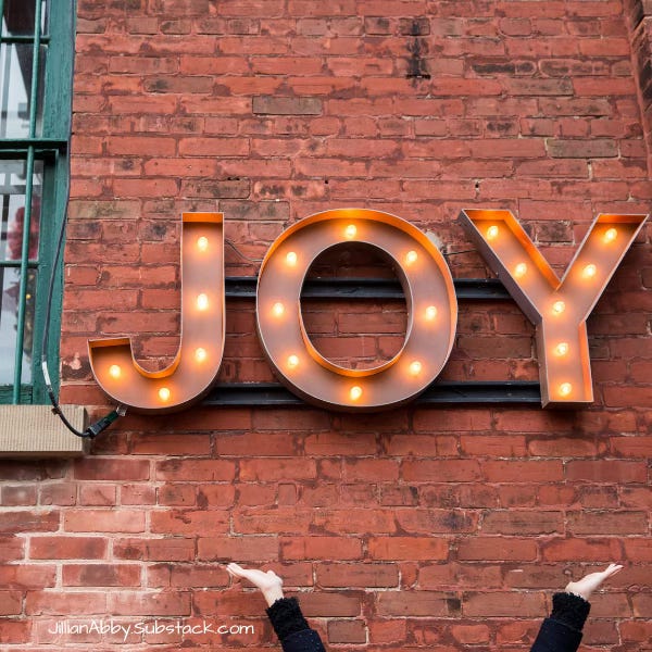 Large marquee-style letters spelling "JOY" with warm light bulbs are mounted on a red brick wall. Below the sign, a person’s arms are raised upward, reaching toward the word. The window of a building is partially visible on the left, and the text “JillianAbby.Substack.com” appears in the bottom left corner.