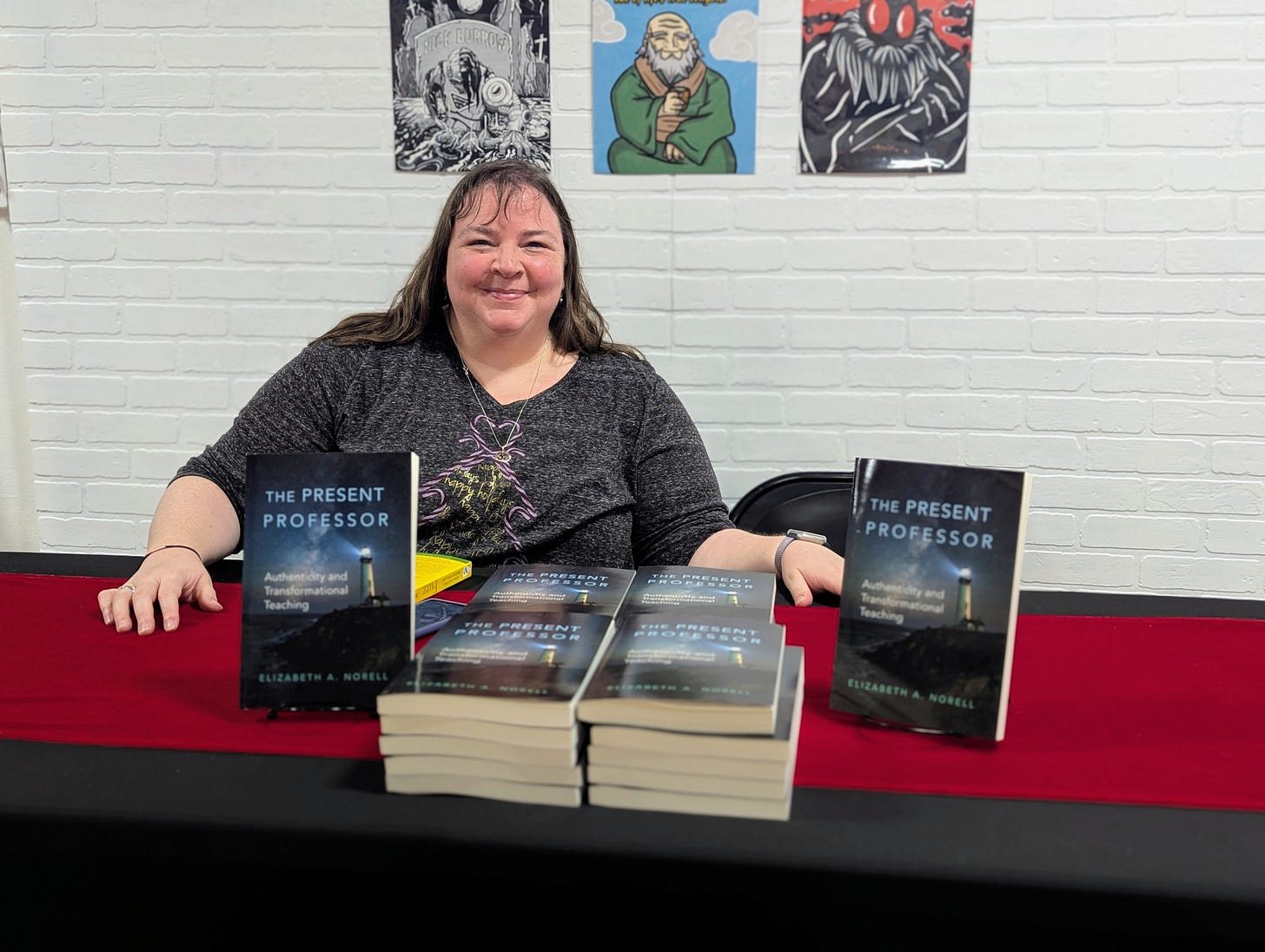 The author is a white woman with a round-ish face and mid-length brown hair. She wears a holiday-themed gray t-shirt with a Christmas tree made out of purple striped candy canes. On the table in front of her are multiple copies of her book, The Present Professor. Behind her are various art prints. The author is a white woman with a round-ish face and mid-length brown hair. She wears a holiday-themed gray t-shirt with a Christmas tree made out of purple striped candy canes. On the table in front of her are multiple copies of her book, The Present Professor. Behind her are various art prints.