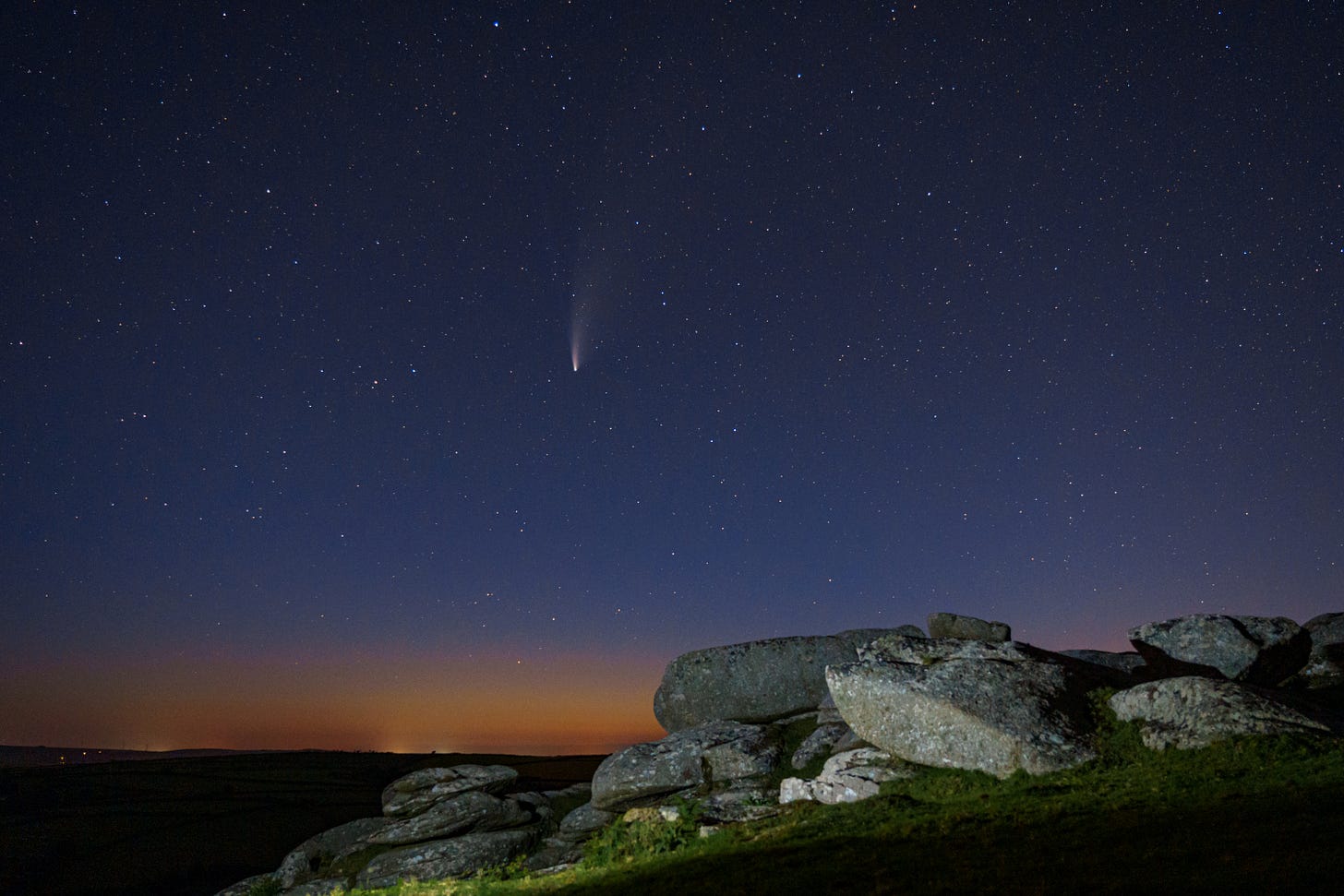 Comet NEOWISE lighting up the northern sky shortly after an orange sunset in Cornwall, UK. In the foreground, the rocks of Helman Tor and light painted.