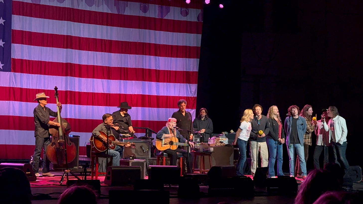 Willie Nelson, band and guests perform in front of a large American flag backdrop.