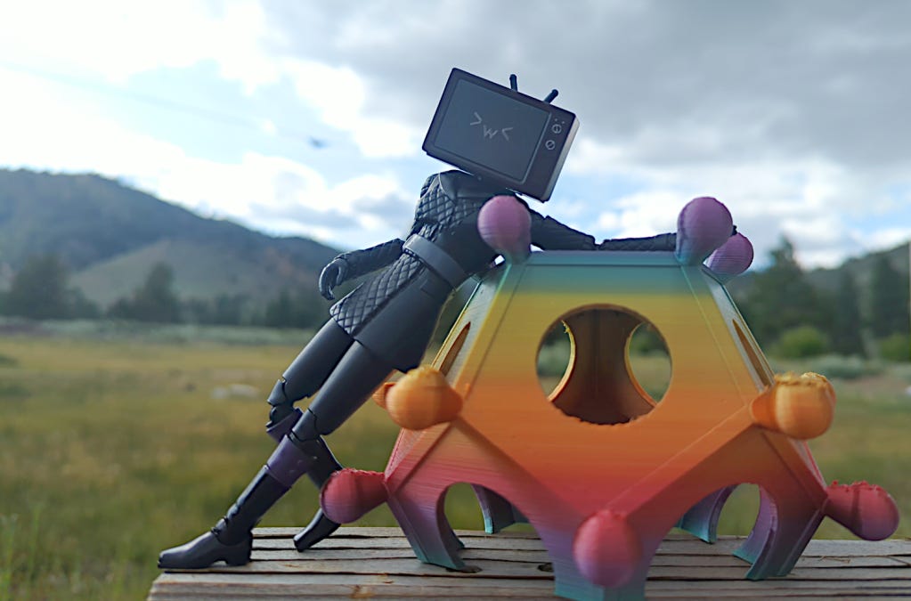A photo of an action figure of the TVhead woman from Skibidi Toilet, going >w<, over a beautiful mountain backdrop. She's reclining against a partially-completed rainbow 3D print of a Roman dodecahedron. Hey, look, it's a blog callback!
