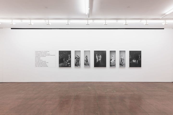 1) Four black-and-white photographs are displayed on a white gallery wall. Each image features people with speech bubbles, conveying a narrative tone in a comic style. These are the images from Tessa's sailor series. Each work is a square, in a thin white frame. The floor is a dark brown tiled flooring. 2) A minimalist art gallery with a white wall displaying seven black and white photos in a row. To the left, the wall features text about The Knight Move series that is displayed to the right of it. Bright overhead lighting. The flooring is a dark brown tile flooring. The beginning, middle, and end photos are large squares, while the 4 images, 2 on either side of the middle work are thin rectangular images.
