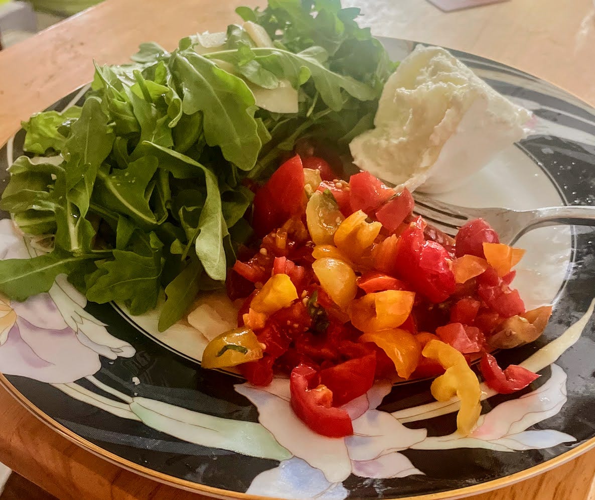 Roughly chopped red and yellow cherry tomatoes, a big pile of bright green arugula and homegrown basil on top of a fresh slice of bruschetta (not visible under the greenery) and a big ball of fresh mozzarella on a black and white floral illustrated plate.