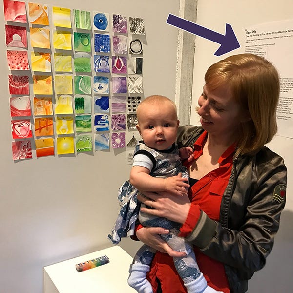 A photo of me holding my baby in front of One Tiny Painting a Day Seven Days a Week for Seven Weeks, made in 2016 and the impetus for Rainbow Squared. This image has an arrow pointing to the wall text behind my head.