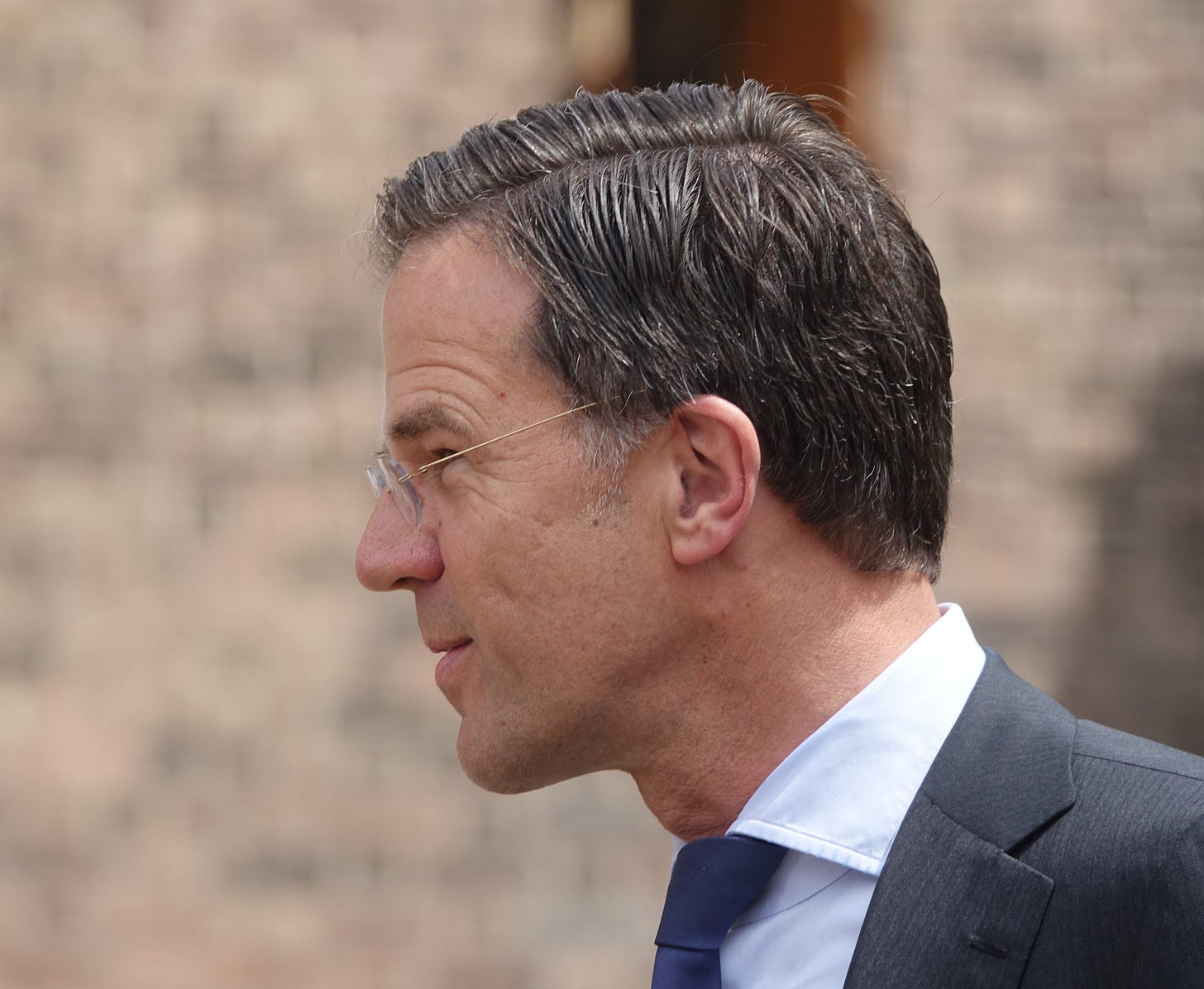 Side profile of NATO Secretary General Mark Rutte wearing a navy suit and blue tie, photographed outdoors with a blurred brick wall in the background.
