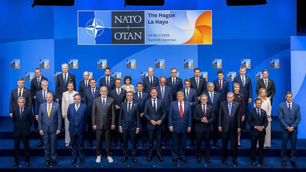 A large group of men wearing suits and four women stand in three rows for a group photos. They are against a blue background and a sign which says 'Nato'