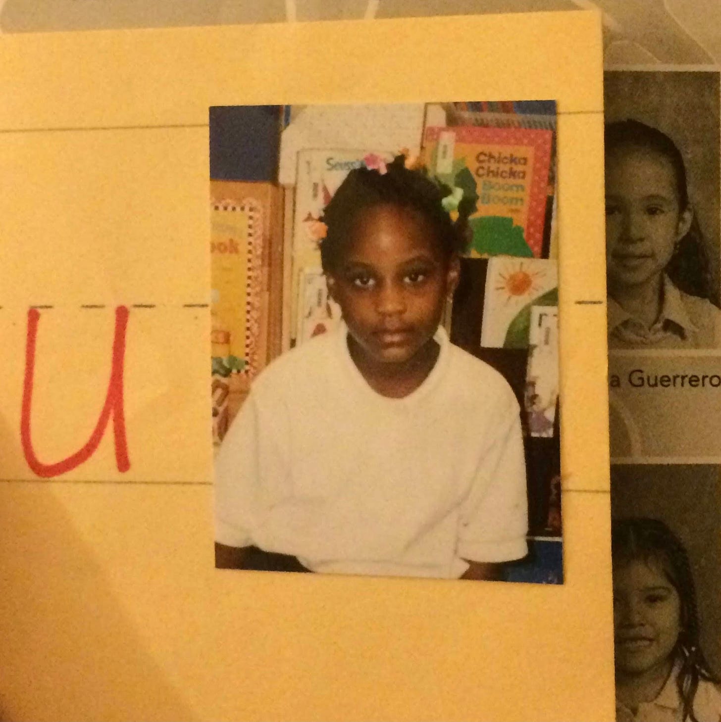 this is a photo of me as a little Black girl with bows clipped in my plaits and a white peter pan shirt as my school uniform top. in the background are children books with the novel chicka chicka boom's cover showing.
