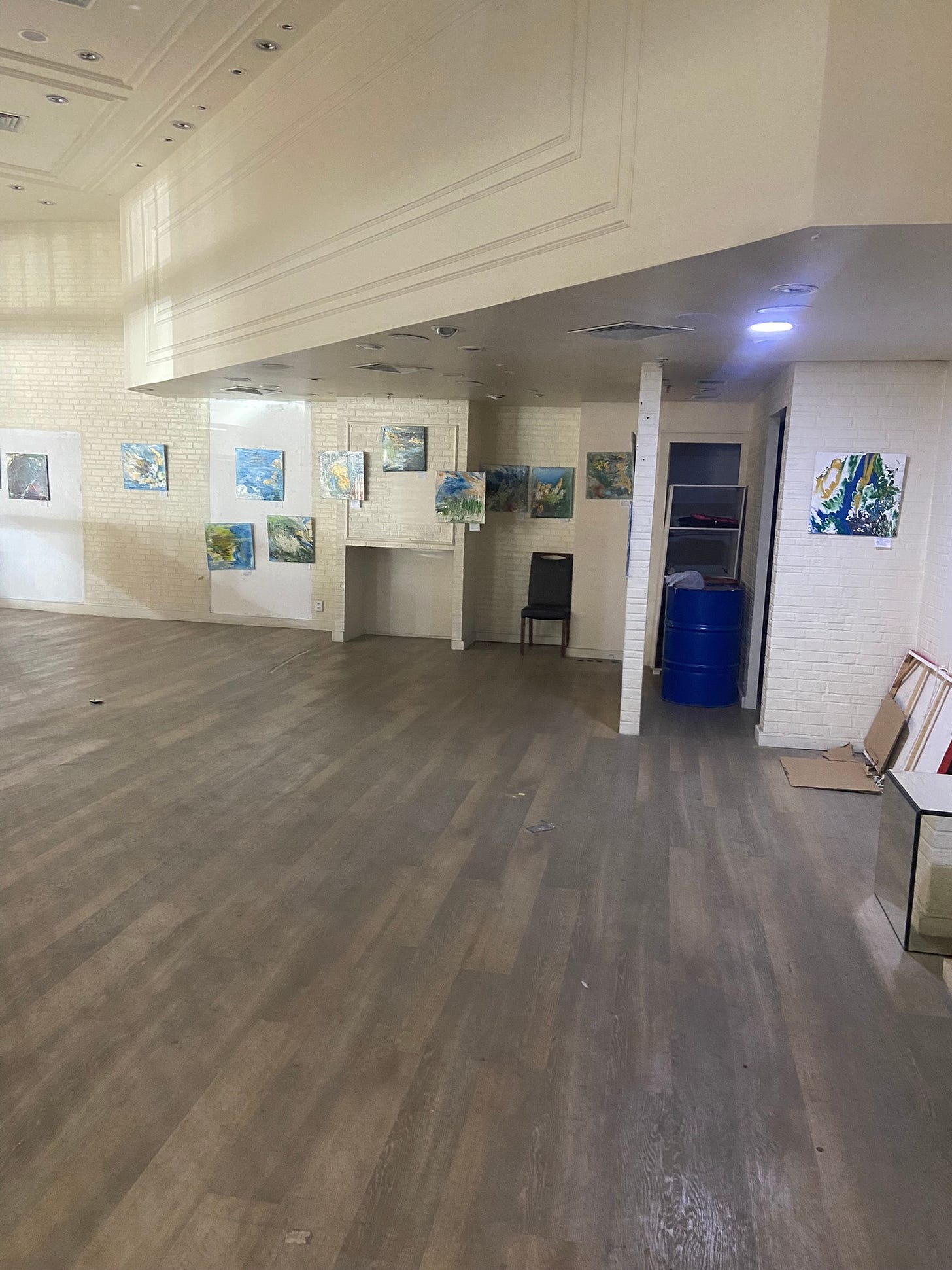galeria com quadros abstratos e coloridos pendurados. pé direito alto em gesso. paredes de tijolo branco. chão de madeira. uma cadeira solitária e um barril azul podem ser observados ao fundo.
