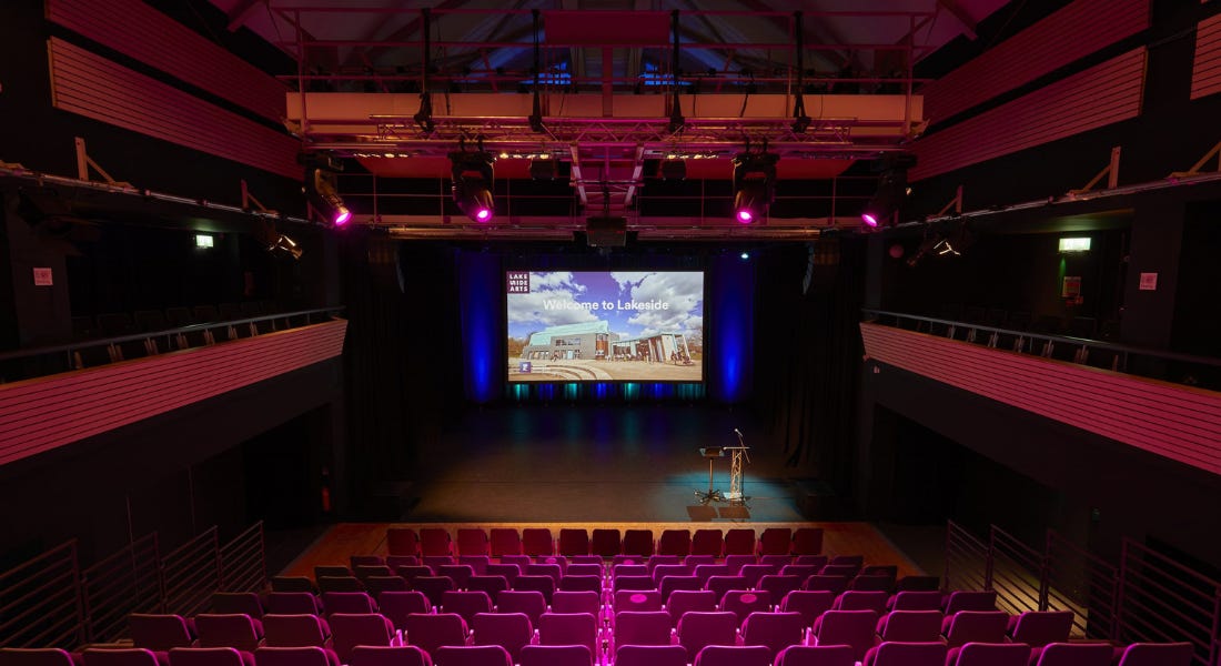 A theatre interior with pink seats, a balcony, and a lit up screen on the stage A theatre interior with pink seats, a balcony, and a lit up screen on the stage