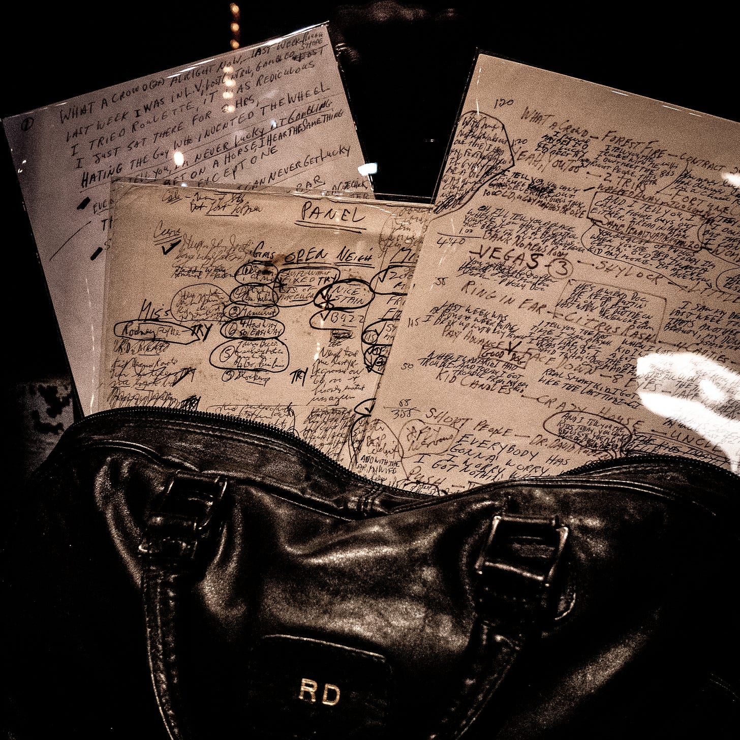 Sepia-tinted photograph of a dark-colored leather bag, monogrammed "RD." Zippered closure of the bag is open, and several pages of handwritten notes are arrenged as if spilling from inside. Most of the notes are single words or phrases -- some the germs of jokes, some apparently Dangerfield's notes to himself: "What a crowd," "Forest fire," "TRY," "Nice stain," "MISS," etc. The only complete (and completely legible) joke says, "Last week I was in LV [Las Vegas]... I tried roulette, it was ridiculous. I just sat there for 2 hours, hating the guy who invented the wheel."