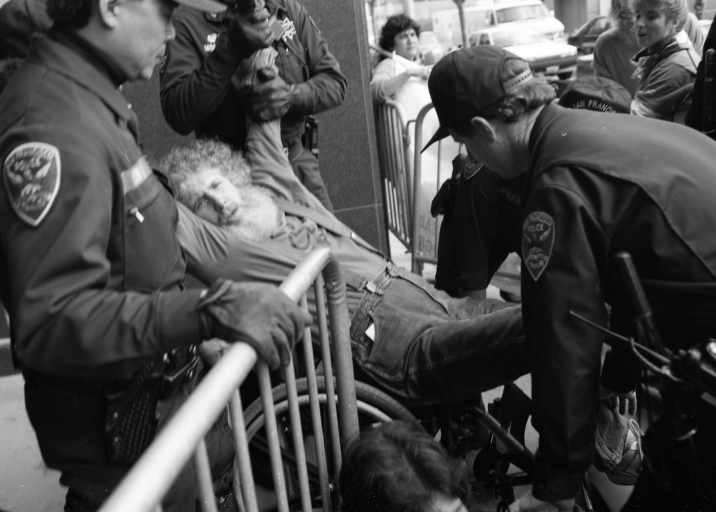 Bob Kafka, an older white man who is a wheelchair user and has curly white hair and a long white beard, being forcibly moved by 3 cops at a barricade.