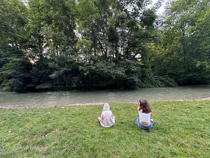 Two teen girls sitting on grass in front of a small river with trees behind it. Two teen girls sitting on grass in front of a small river with trees behind it.
