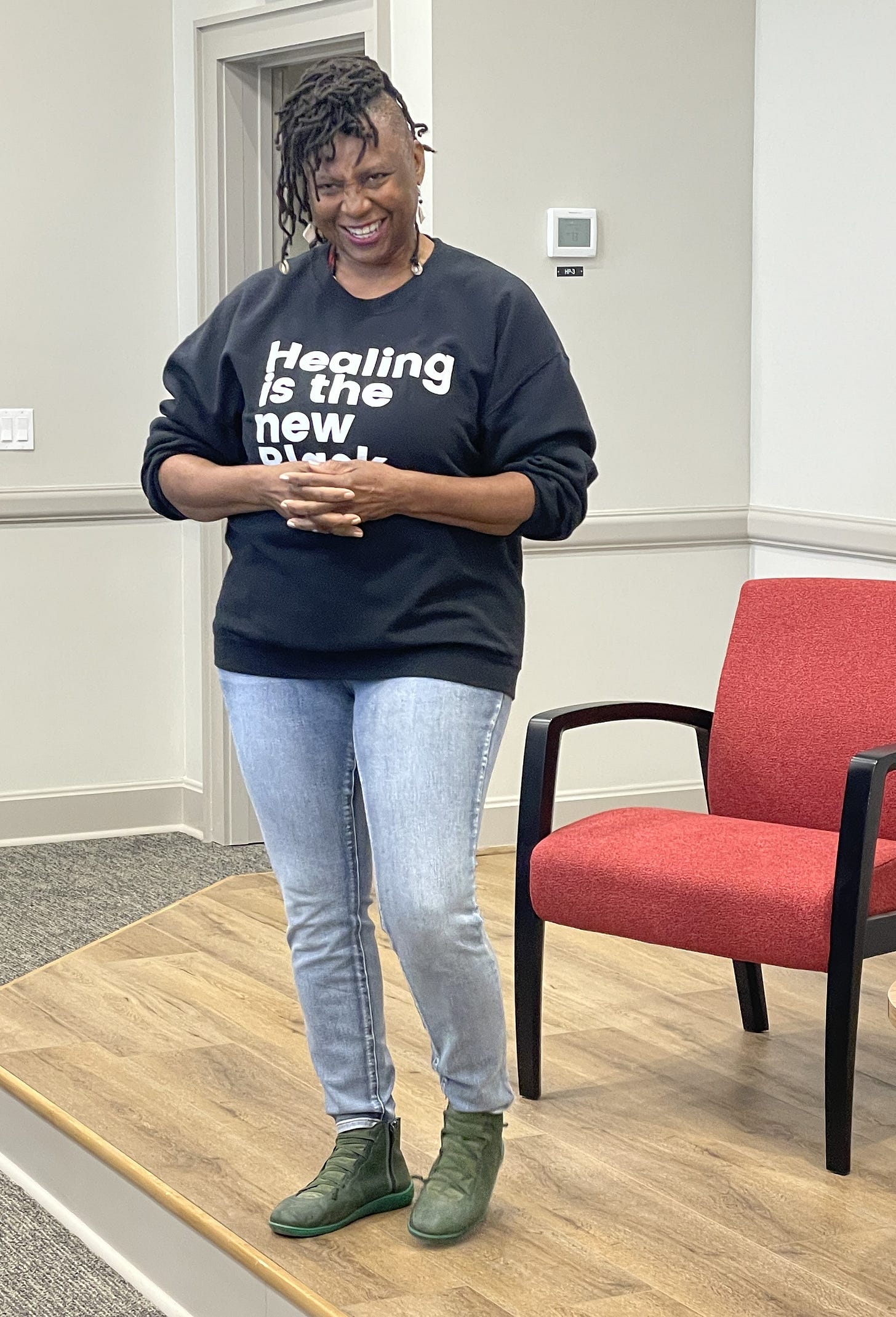 A black woman with locs gathered atop her head and wearing a black sweatshirt and jeans, smiles as she addresses a crowd.