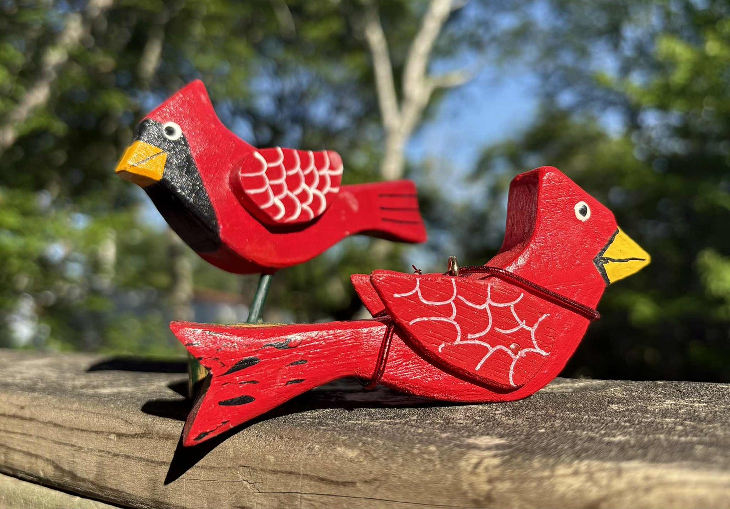 Two wooden ornaments carved and painted as male northern cardinals of red sit on a worn wooden railing overlooking a backyard of trees.