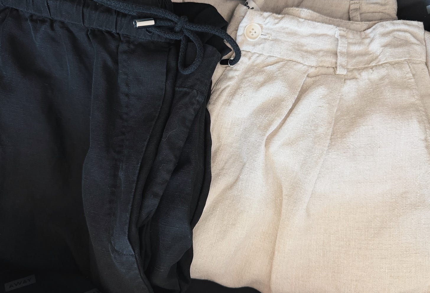 Cream linen trousers and black linen-blend drawstring trousers arranged side by side in an open suitcase.