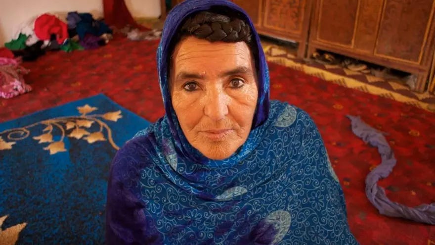 Una donna con un foulard blu scuro e un foulard a motivi geometrici blu chiaro con decorazioni bianche guarda in avanti. I suoi capelli scuri sono intrecciati e visibili sopra la fronte. Ha gli occhi castani con palpebre pesanti e alcune rughe intorno a essi. Ha un naso dritto e labbra sottili. Lo sfondo è un tappeto rosso a motivi geometrici con un piccolo tappeto blu sotto di lei sulla sinistra. 