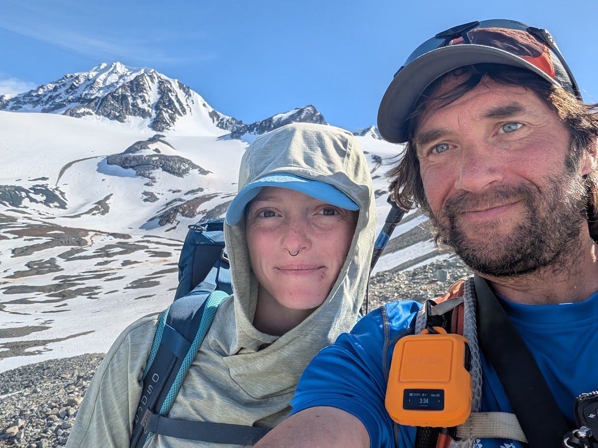 Two hikers pose close to the camera, one in a pale hood and cap with a nose ring, the other in an orange cap and blue shirt with a sunburned face and an orange satellite messenger clipped to his chest. Behind them a snow-covered peak and glacier basin rise under a bright blue sky.