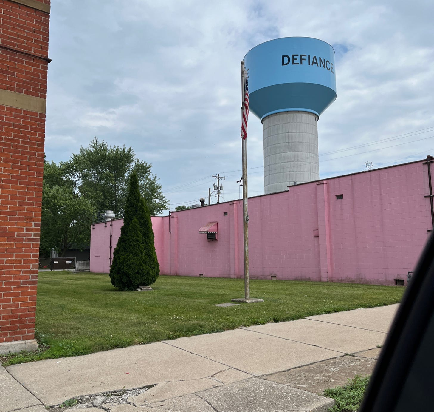 A water tower is shown against a gray sky, in front of a concrete block building painted pink. The water tower says DEFIANCE.
