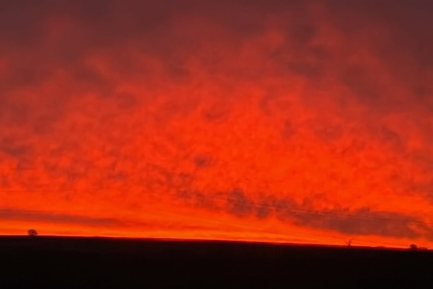 A vivid sunset fills the sky with fiery shades of red and orange, fading into deep purple at the top. The horizon is a dark silhouette of rolling land with a few small trees, creating a striking contrast against the glowing sky.