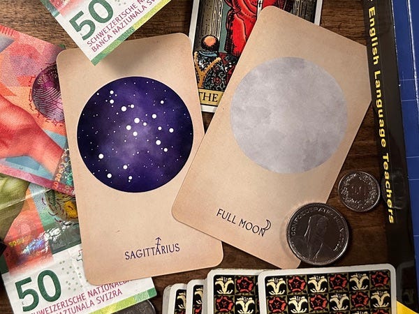 Two cards are on a wooden table. The one on the left says Sagittarius. The one on the right says full moon. Paper money, coins, a feather and a book surround them.