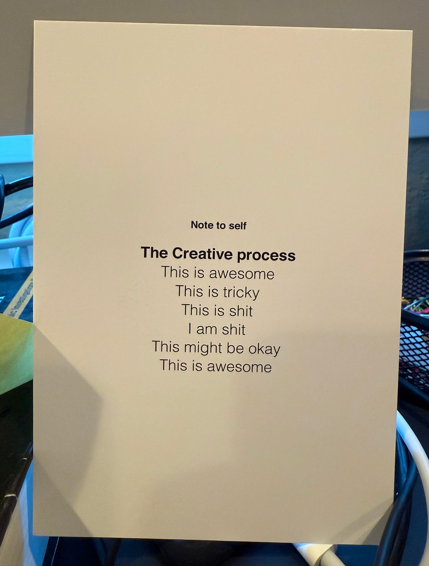 A printed card titled 'Note to self' with the heading 'The Creative process,' listing the emotional stages: 'This is awesome, This is tricky, This is shit, I am shit, This might be okay, This is awesome.' The card is propped up on a cluttered desk with cables and office supplies visible in the background. A printed card titled 'Note to self' with the heading 'The Creative process,' listing the emotional stages: 'This is awesome, This is tricky, This is shit, I am shit, This might be okay, This is awesome.' The card is propped up on a cluttered desk with cables and office supplies visible in the background.