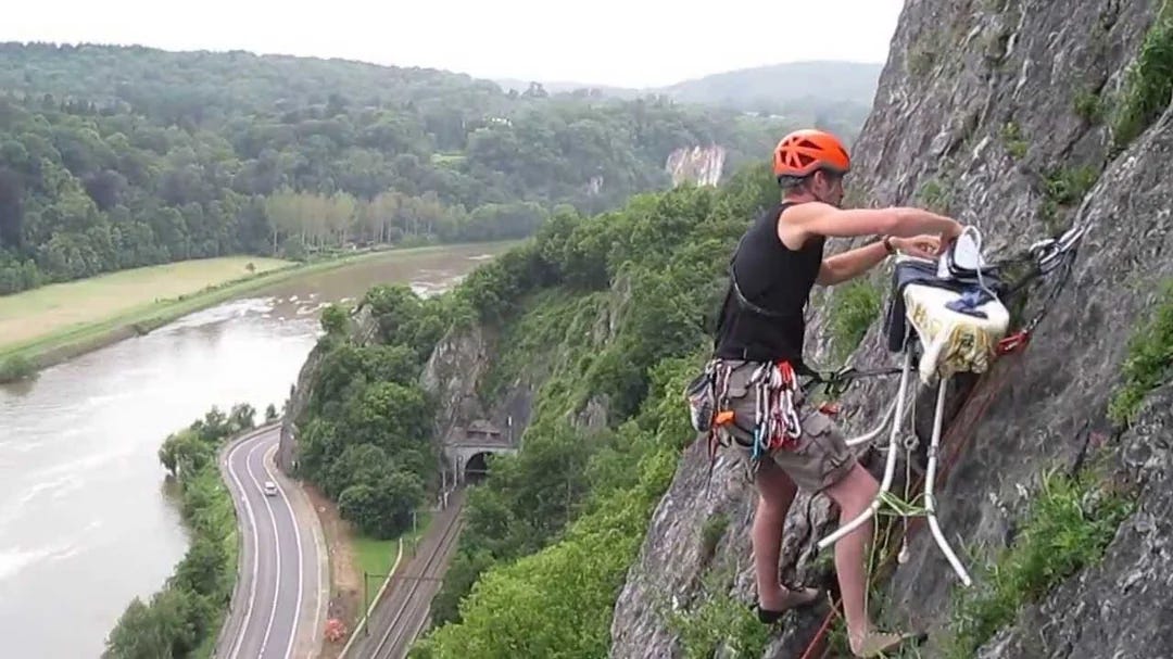 Rock climber in orange helmet scales a cliff while ironing. Verdant landscape and winding river with roads in the background.