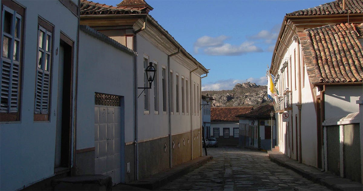Fotografia de uma rua estreita e calçada com pedras, em um centro histórico. Dos dois lados, há casarões coloniais de um ou dois andares, pintados principalmente de branco Fotografia de uma rua estreita e calçada com pedras, em um centro histórico. Dos dois lados, há casarões coloniais de um ou dois andares, pintados principalmente de branco