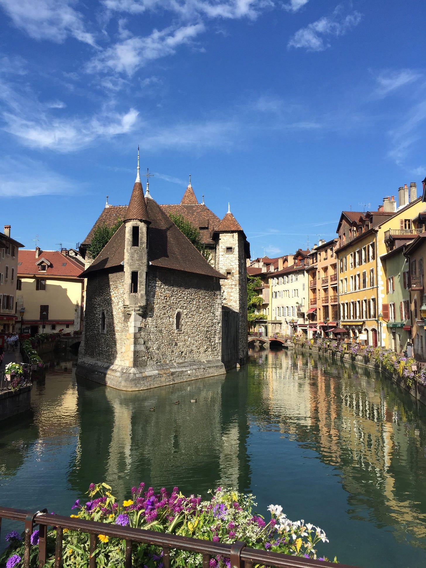 The Palais de l'Île in Annecy, France, reflected in the canal, with colourful buildings, flower-filled balconies, and a bright blue sky.