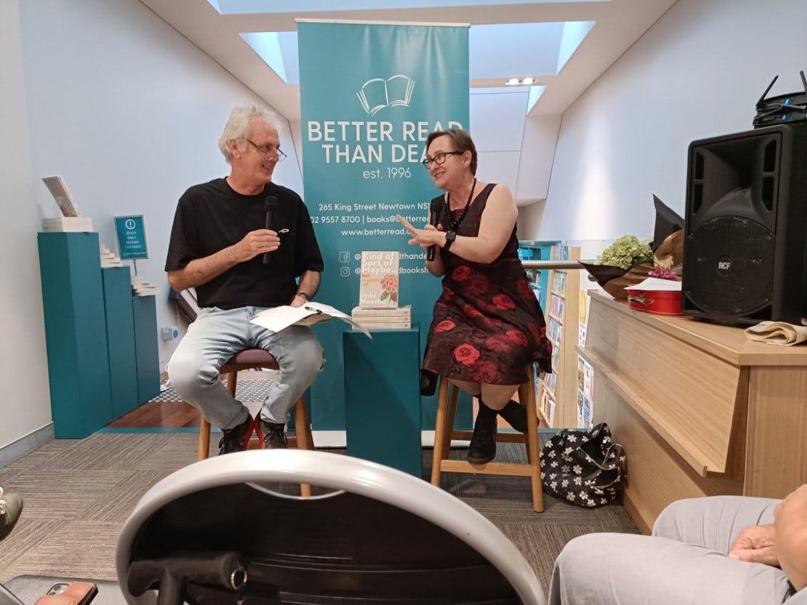 A man and woman sit on stools, "in conversation" with microphones. The woman looks like she is telling the man a HILARIOUS anecdote. The man looks amused or perhaps a little scared. 