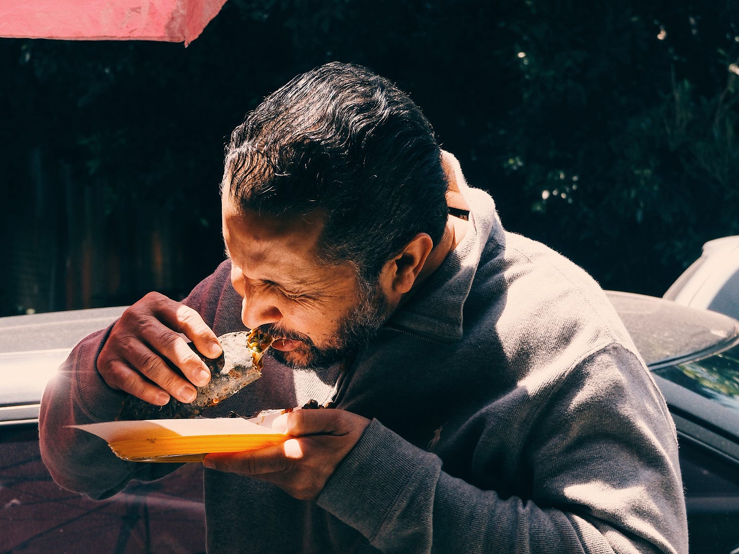 A man eats a taco from a yellow plate while standing on a street in Mexico City, with a pink umbrella and parked car behind him.