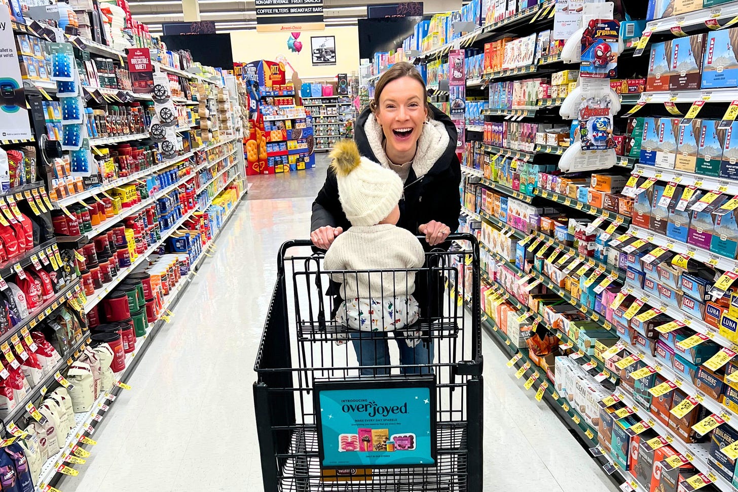 T1D health coach and mom Allee Peterson smiling while grocery shopping with her young child sitting in the cart in a brightly lit grocery aisle.