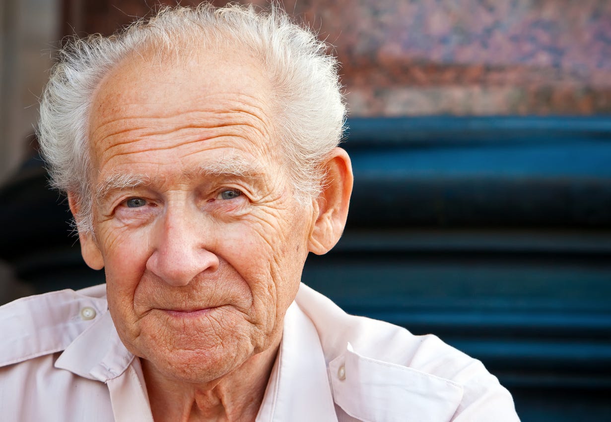 White, aging man with white hair and no glasses with a smirk on his face as he looks into camera.