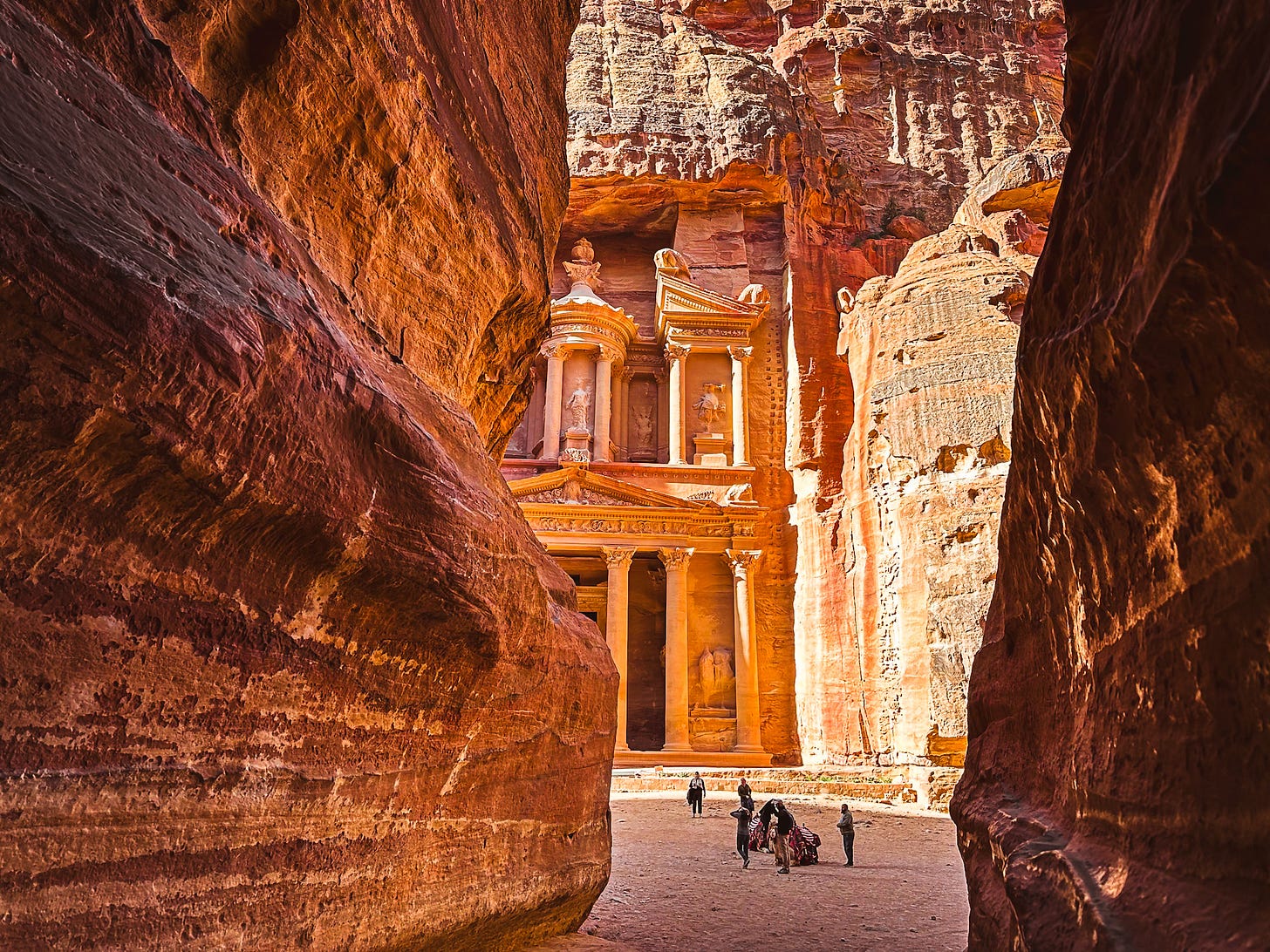 High reddish rock walls obscure the right and left sides of the image, while sunlight shines on a building carved into the rock visible from the narrow channel between the rock walls.