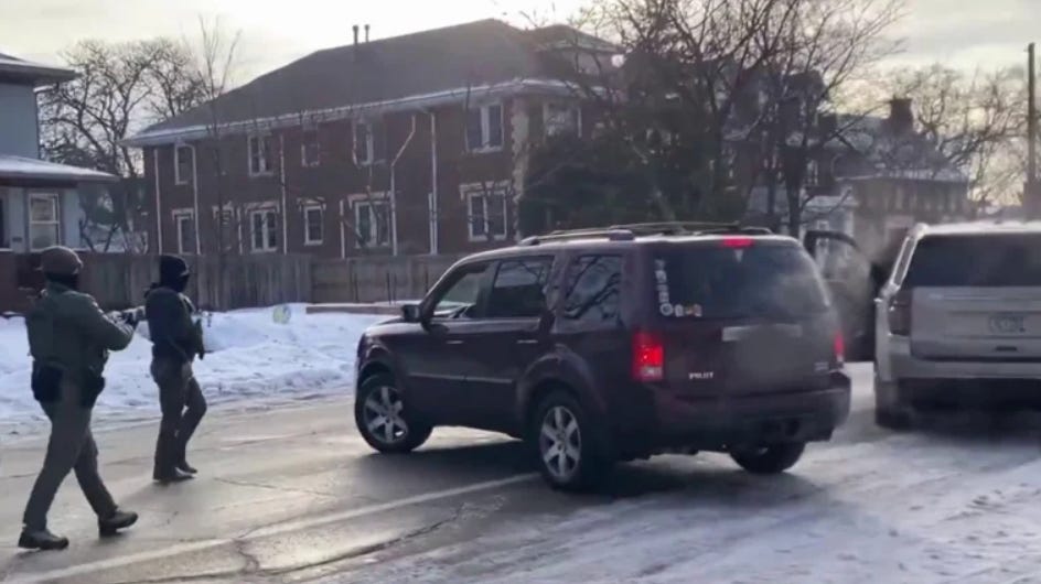 Two armed agents in tactical gear approach a dark SUV on a snowy street, with another vehicle nearby. Two armed agents in tactical gear approach a dark SUV on a snowy street, with another vehicle nearby.
