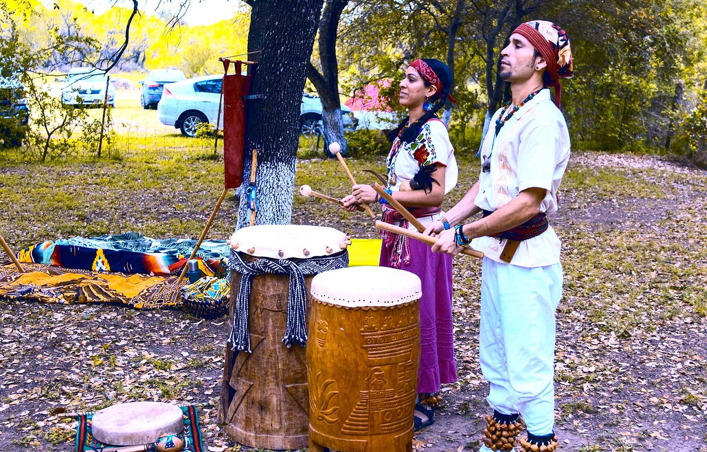 Gera and his wife Vanessa are standing in a wooded clearing, dressed in white. They are playing large ceremonial drums.