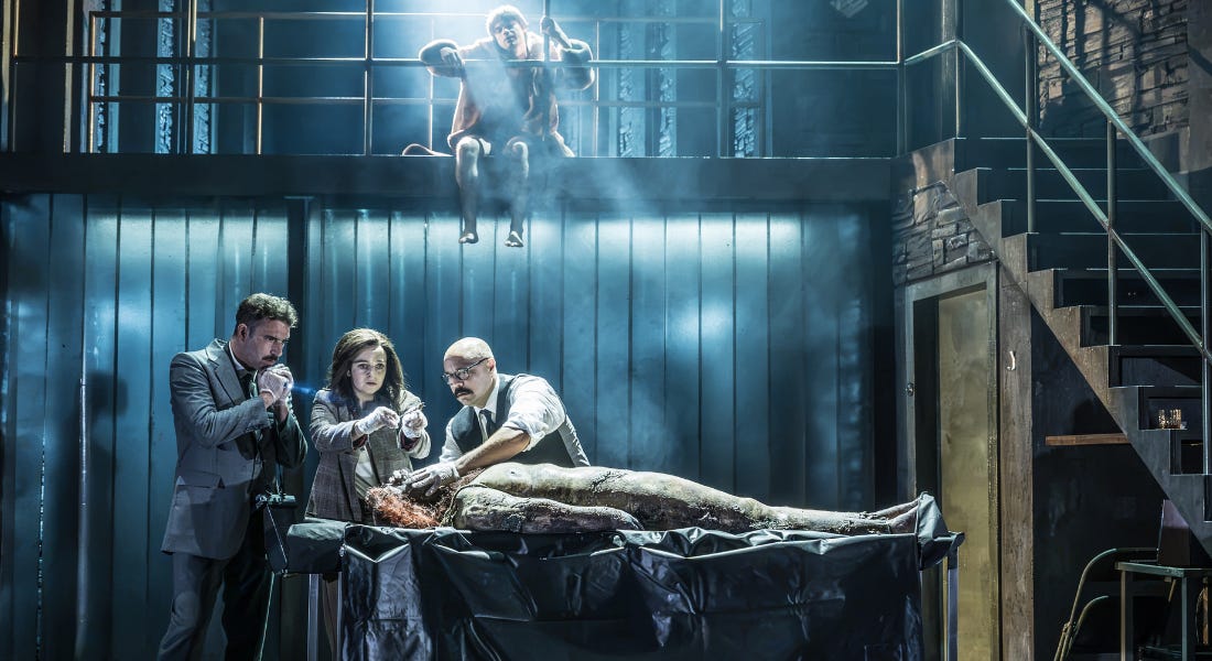 A stage set with three people around an autopsy table, with a person sitting on a ledge above them A stage set with three people around an autopsy table, with a person sitting on a ledge above them