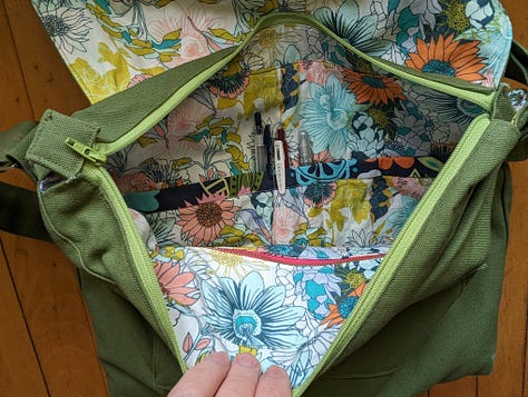 Five photos of the Mythical Messenger Bag. In the first, I (a white lady, aged 52) am standing in the hallway wearing the bag. The other photos are of the bag on its own. The bag is olive green canvas with an adjustable strap. There is hot pink decorative stitching. There is an exterior welt pocket on the back with a hot pink zipper. There is a pocket on the front with a blue square patch that says "Make Art" in a red cursive font. The inner lining fabric is floral -- blue, green, coral line drawn flowers on a white background. Inside the bag, there are two pockets. One is a zippered pocket with a hot pink zipper. The other is a divided pocket with slots for pens, a phone, and something else roughly cell-phone-sized.