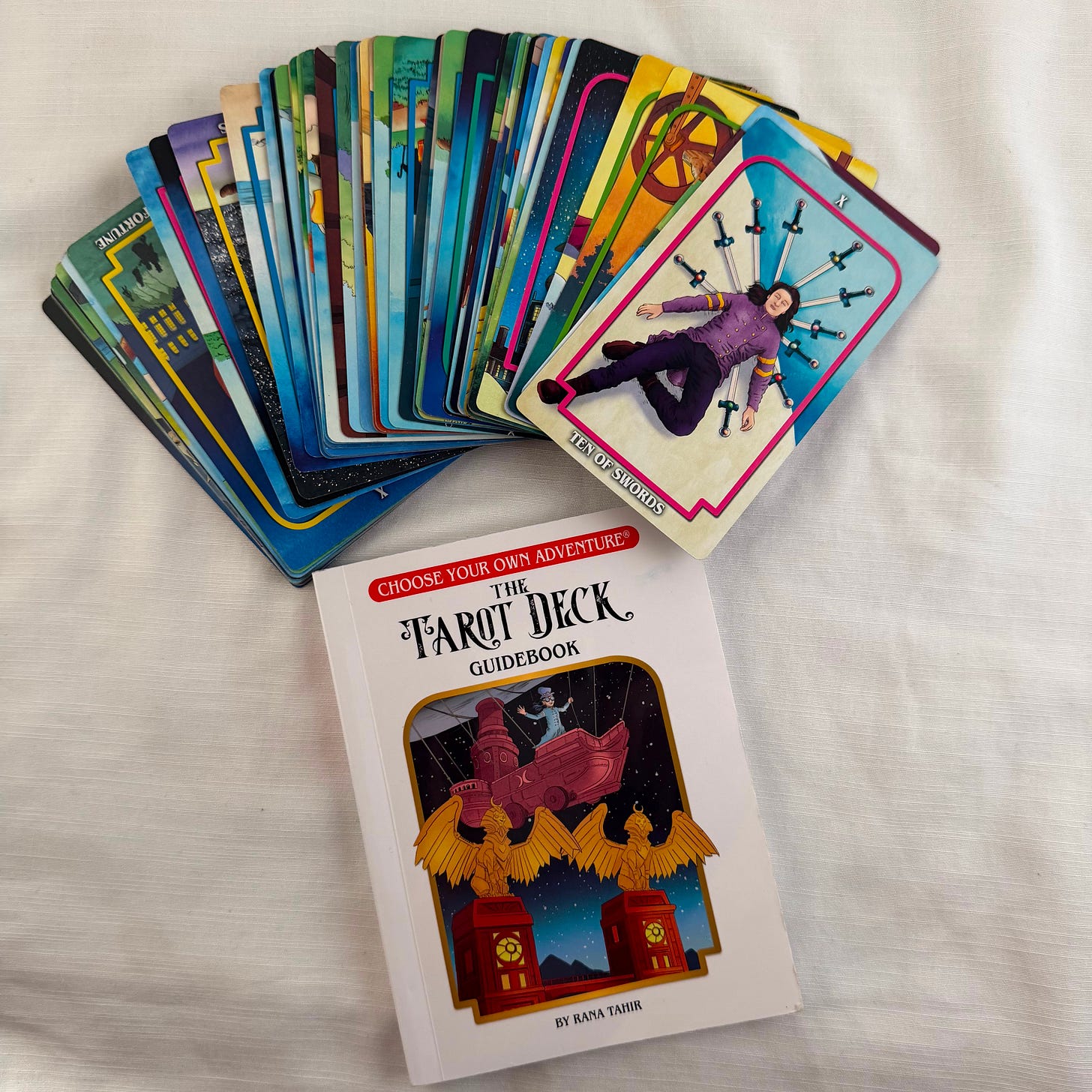 The Choose Your Own Adventure Tarot Deck fanned out on a white linen backdrop, with the booklet beside it. The top card of the fan shows the Eight of Swords — a figure in purple lying beneath a row of swords against a pale blue background. The booklet cover features Choose Your Own Adventure's signature red and white logo at the top, the title "The Tarot Deck Guidebook" in stylized lettering, and an illustration of a winged figure piloting a small airship between two glowing towers under a starry sky. By Rana Tahir. The Choose Your Own Adventure Tarot Deck fanned out on a white linen backdrop, with the booklet beside it. The top card of the fan shows the Eight of Swords — a figure in purple lying beneath a row of swords against a pale blue background. The booklet cover features Choose Your Own Adventure's signature red and white logo at the top, the title "The Tarot Deck Guidebook" in stylized lettering, and an illustration of a winged figure piloting a small airship between two glowing towers under a starry sky. By Rana Tahir.