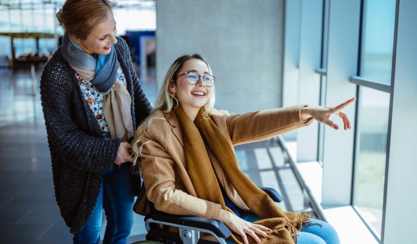 A woman in a wheelchair smiles and points excitedly toward something outside a large window, while another woman stands behind her, gently pushing the chair and sharing the moment. They appear to be in a bright, modern building such as an airport or station, with sunlight streaming through the glass and a warm, joyful atmosphere between them.