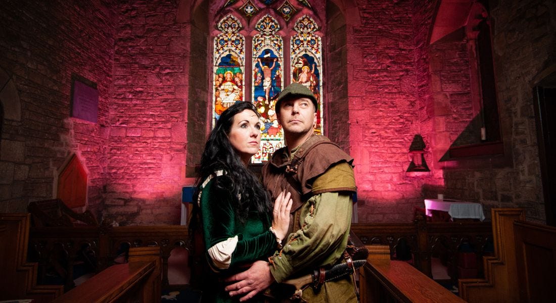 Two people dressed as Maid Marian and Robin Hood standing in the middle of a brick church with pink lights and a stained glass window behind them